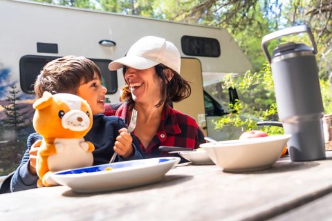 Mother and son laughing together while enjoying breakfast at a picnic table outside their camper