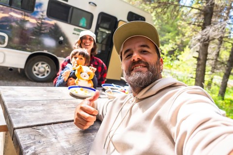 Happy family taking a selfie while having lunch on a wooden table near their camper van in the
