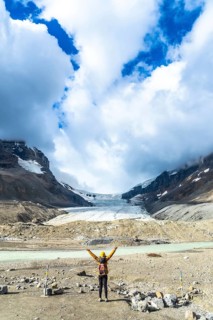 Female hiker with backpack and yellow jacket raising arms in front of the athabasca glacier, in the
