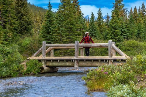 Female hiker wearing red plaid shirt and white hat is leaning on a wooden bridge railing over a