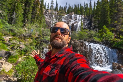 Tourist wearing sunglasses and red and black shirt taking selfie with outstretched arm in front of