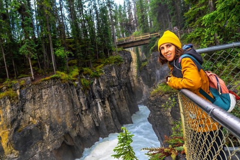Female tourist in a yellow jacket and beanie leaning on a metal railing, admiring the stunning view