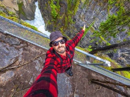 Photographer wearing sunglasses and a bucket hat takes a selfie at sunwapta falls in jasper