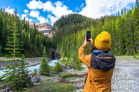 Hiker capturing smartphone images of a turquoise waterfall flowing through a lush pine forest along