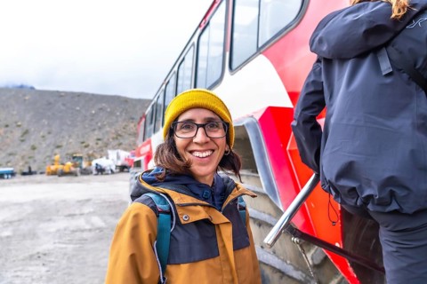 Tourist wearing a yellow beanie and brown jacket smiles near a large red bus at the athabasca