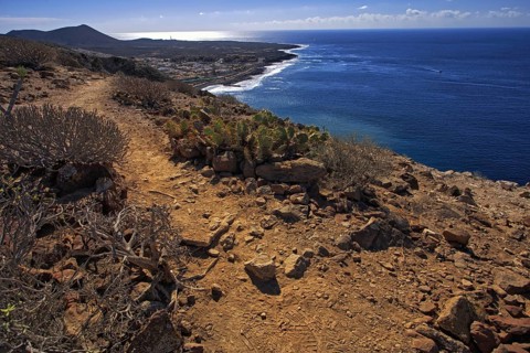 A rocky path leads along the coast, the sea shines in the distance under a clear sky, Los