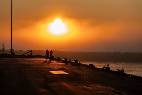 Silhouettes of people fishing at harbour at sunset under a warm orange sky, Vardø, Finnmark, Norway