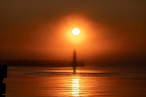 A lighthouse at the Vardö pier stands silhouetted against an orange sunset over the sea reflecting