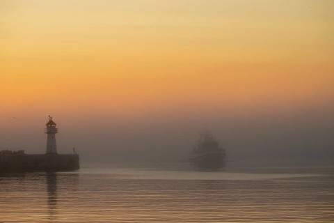 A lighthouse at the entrance to Vardö harbour at sunset with a ship in fog on calm water, Vardø,
