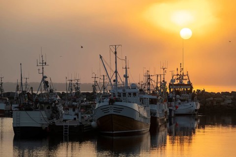 Fishing boats are moored in the harbor at sunset, the sea reflecting the warm colors of the sky,