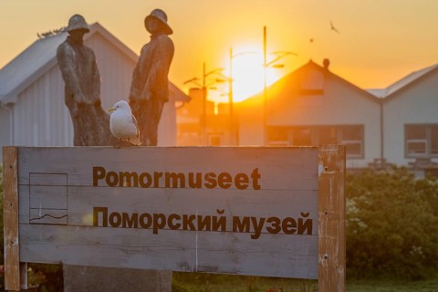 A seagull sits on a museum sign in Norwegian and Russian in front of sculptures and wooden