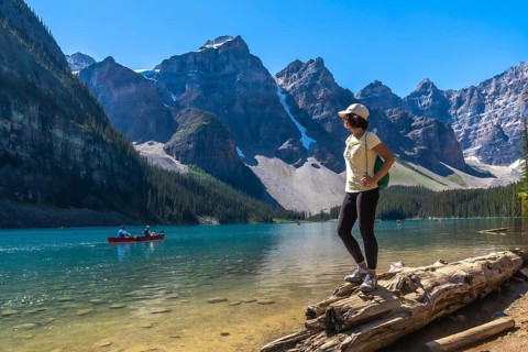 Female tourist standing on a log, admiring turquoise waters of moraine lake with canoers,