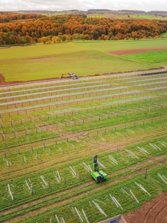 Tractor works in a green field with solar plant construction beginning, surrounded by autumnal