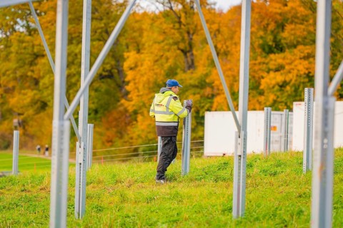 A worker wearing protective clothing erects metal structures on a grass-covered area in autumn,