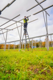 Man on a ladder assembles a metal structure on an open green area, Energiewende, construction of PV