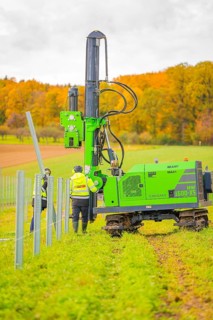 Two workers fix poles with a large, green machine in a field in an autumn landscape, Energiewende,
