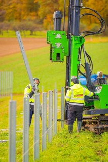 Workers install metal poles in a field with a large green machine in the background, Energiewende,