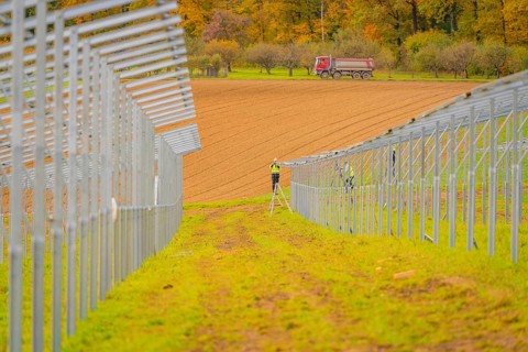 A farm worker on a ladder between metal structures with a tractor in the background in an autumn
