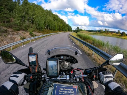 A motorcyclist's view of an empty country road surrounded by green trees and a lake under a blue