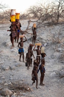 Himba woman and children run to fetch water with canisters through dry countryside, traditional