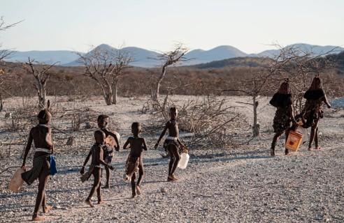Himba woman and children run to fetch water with canisters through dry countryside, traditional