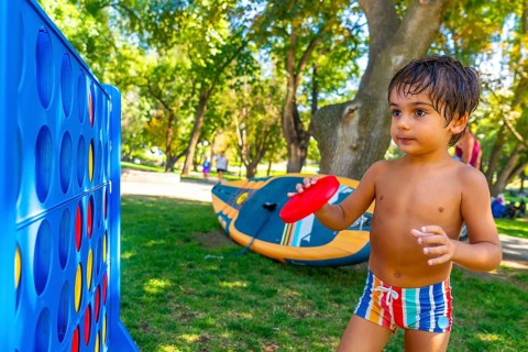 Young child holds a red flying disc, standing near a large connect four game in a park in kamloops,