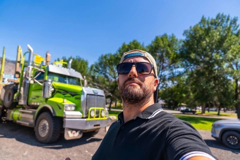 Trucker wearing sunglasses and baseball cap taking selfie with his logging truck in a parking lot