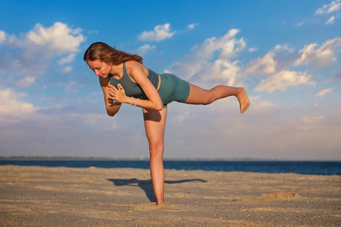 A woman performs Virabhadrasana Warrior 3 pose on the beach during sunset. Her strong stance and