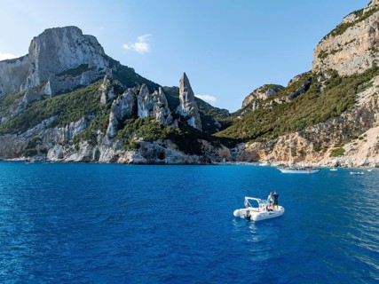 Couple on motorboat off picturesque rocky coast, cliffs with L'Aguglia pinnacle, blue sea and Cala