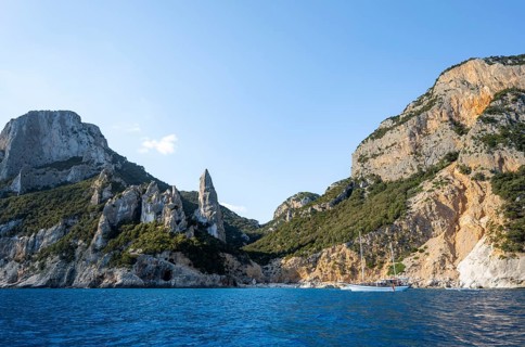 Excursion boat off picturesque rocky coast, cliffs in morning light, L'Aguglia pinnacle, blue sea