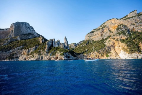 Excursion boat off picturesque rocky coast, cliffs with caves in morning light, L'Aguglia rock pin,