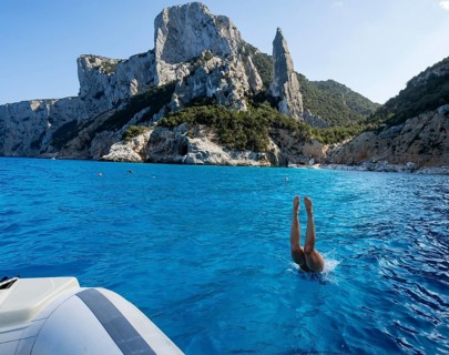 Young woman jumping from a boat into the water, picturesque rocky coast, cliffs with L'Aguglia rock