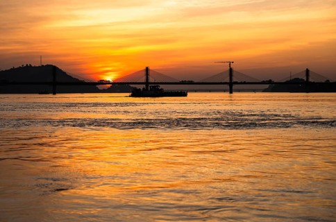 A boat sails on the river Brahmaputra during sunset, with the silhouette of a cable-stayed bridge