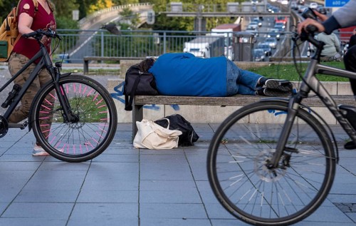 Man sleeping on a bench, travelling bag serves as pillow, in the city centre, passers-by walk by