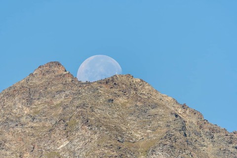Majestic mountain peak stands tall against vibrant blue sky as large moon rises just behind it. The