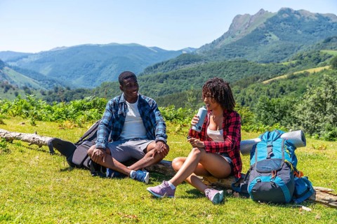 Two hikers are taking a break during their mountain trek, sitting on a log, drinking water and