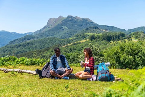 Two hikers are taking a break, sitting on grassy mountaintop and enjoying refreshing drinks with