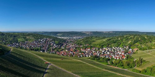 Panorama of a small village view surrounded by vineyards and green hills under a clear sky,