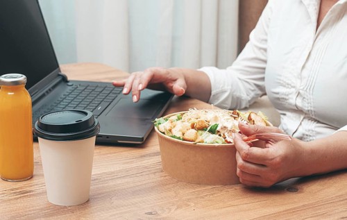 A woman is working on a laptop at home, there is food on the table, salad, coffee and juice,