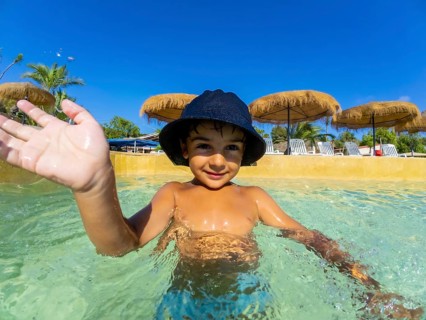 Smiling child in a sun hat waves while playing in a clear swimming pool, surrounded by straw