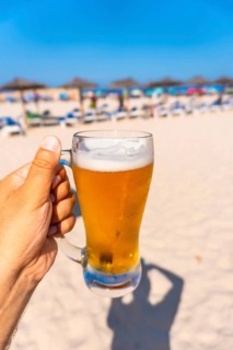 Hand holding glass of cold beer on beach with blurred beach umbrellas and sunbeds in background