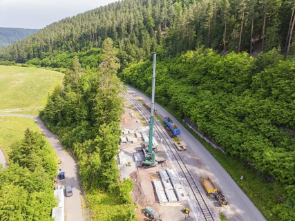 Construction work along a railway line in a green forest area, crane and concrete blocks visible,