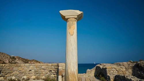 Freestanding stone column in front of a historical site with blue sky in the background, Agios