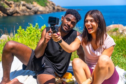 Happy young multi ethnic couple filming with action camera while having picnic by the sea during