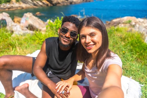 Young multiracial couple holding hands and taking a selfie while enjoying a sunny day by the sea