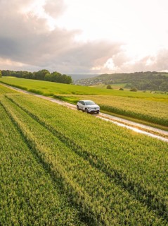 A car drives on a narrow road along green fields under a cloudy sky, Deer E- CarSharing, Renault