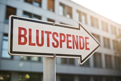 White arrow sign with German text Blutspende (Blood Donation) text in front of sunlit hospital.