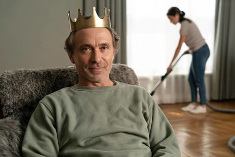 Portrait of middle-aged man with golden crown relaxing on couch while woman vacuums in background.