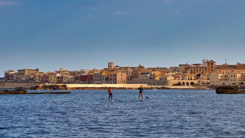 Two paddleboarders on the sea in front of a town with historical buildings under a blue sky, boat
