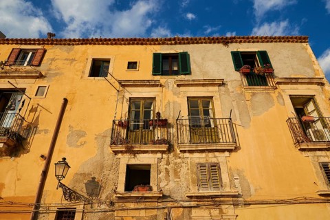 Shows the weather-beaten facade of a building with balconies and windows, blue sky above, Ortigia
